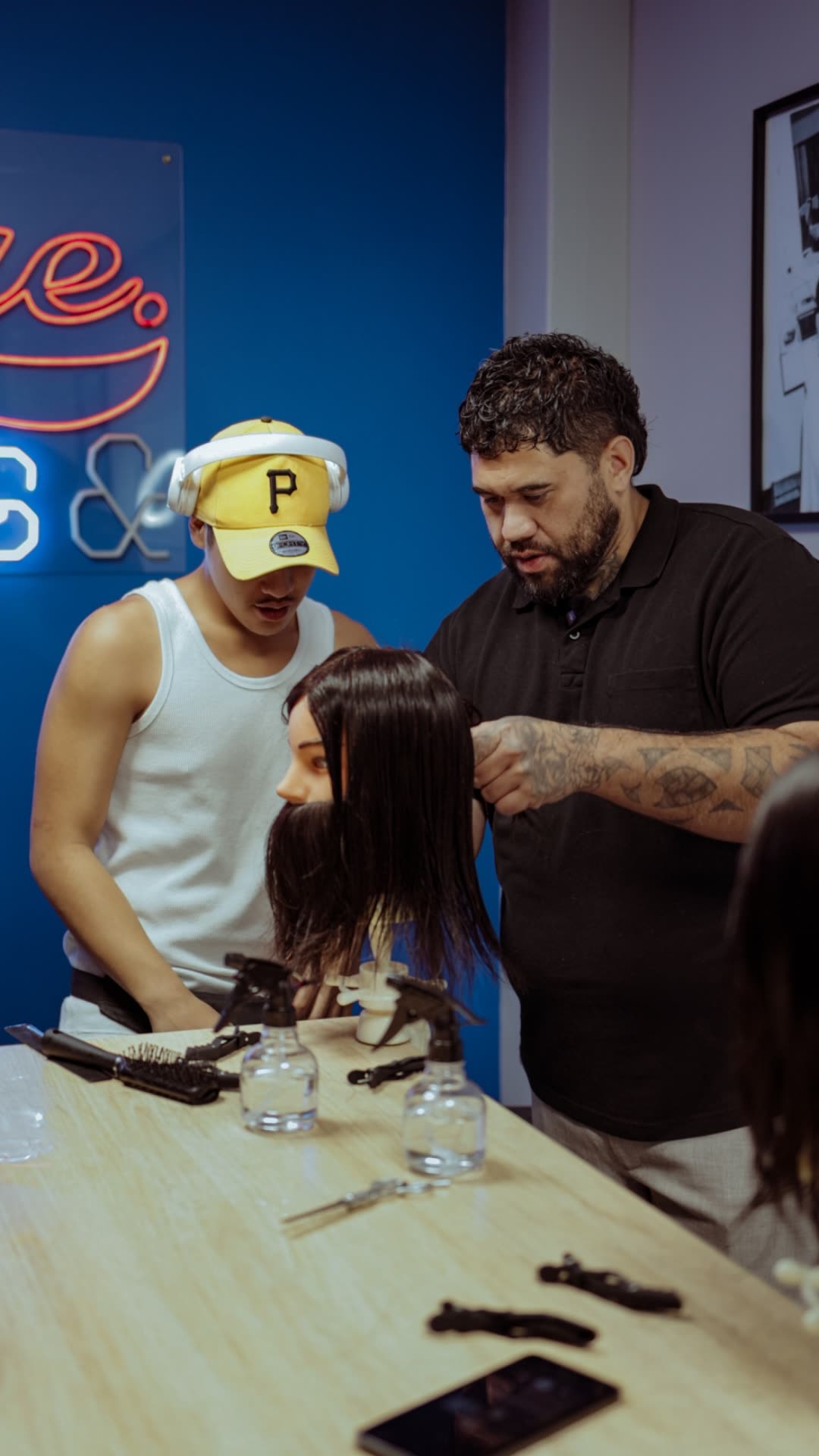 A tutor and student learning barbering tools beside the neon sign
